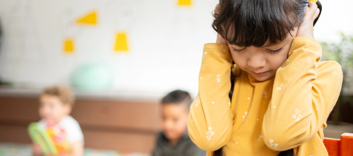 What is sensory overload in autism: child covering ears in a noisy classroom while other children play in the background.